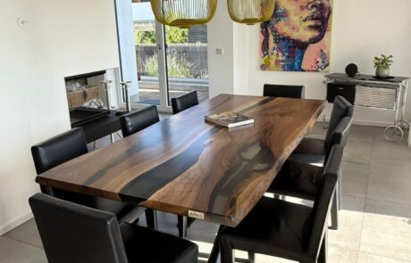 Großer rechteckiger Echtholztisch mit schwarzem Epoxidharz-River und sichtbarer Baumkante steht zentral im Essbereich. Der Tisch ist von schwarzen Lederstühlen umgeben und darüber hängen zwei lange gelbgrüne Pendelleuchten vor einer farbigen Portraitwand.