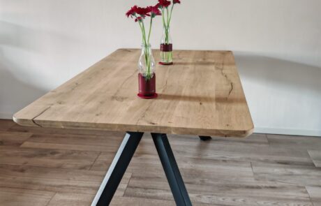 Rechteckiger Massivholztisch aus geölter Eiche mit sichtbarer Maserung und dezenter Naturkante steht auf schwarzem V-Metallgestell. Der Tisch steht in einem hellen Essbereich unter einer schwarzen Halbkugel-Pendelleuchte und ist mit zwei Glasvasen mit roten Blumen dekoriert.