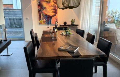 Sechssitziger Massivholztisch mit markanter Naturkante und lebhafter Holzmaserung steht in einem modernen Esszimmer. Schwarze Lederstühle und große Draht-Pendelleuchten ergänzen die helle, luftige Raumgestaltung.