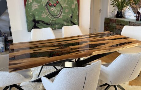 Ein rechteckiger Massivholztisch mit dunklem Epoxidharz-River und natürlicher Baumkante steht auf einem schwarzen Metallkreuzgestell. Der Tisch befindet sich in einem modernen Esszimmer vor einem großen Porträt und ist von weißen Polsterstühlen auf hellem Lammfellteppich umgeben.