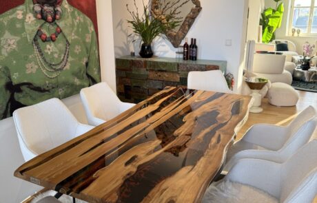 Ein Massivholztisch mit glänzendem schwarzem Epoxidharz-River und sichtbarer Naturkante steht auf einem schwarzen Metallgestell in einem hellen Esszimmer. Der Tisch ist von weißen Polsterstühlen auf einem Fellteppich umgeben; im Hintergrund sind eine dekorative Kommode, ein geschnitzter Spiegel und großformatige Wandkunst zu sehen.