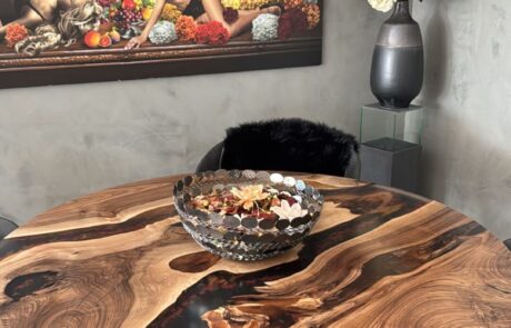 Runder Massivholztisch mit lebendiger Holzmaserung und schwarzen Epoxidharz-Einsätzen steht auf einem schwarzen Metallgestell in einem Essbereich. Auf der Platte liegt eine dekorative Schale, im Hintergrund ist ein großes Wandbild und eine Vase mit Blumen sichtbar.