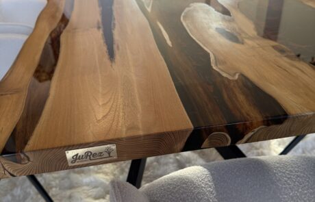 Nahaufnahme eines Massivholztisches mit naturbelassener Baumkante und dunkel eingefärbtem Harz-River auf der Tischplatte. Das Blatt ruht auf einem schwarzen Metallgestell und steht zwischen hellen, gepolsterten Stühlen auf einem flauschigen Teppich.