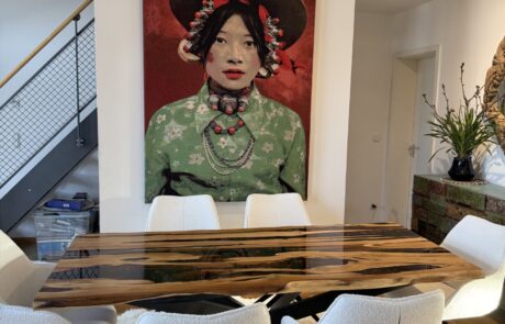 Ein Massivholztisch mit glänzender Platte und dunklen Epoxidharz-Rivern zeigt ausgeprägte Holzmaserungen und sichtbare Naturkanten. Der Tisch steht auf einem schwarzen Metallgestell in einem modernen Essbereich mit weißen Polsterstühlen, Teppich und großformatiger Wandkunst.