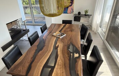Ein rechteckiger Walnuss-Massivholztisch mit schwarzen Epoxidharz-Rivers und sichtbarer Naturkante steht zentral in einem hellen Essbereich. Schwarze Lederstühle und große goldfarbene Pendelleuchten rahmen das exklusive Tischdesign.
