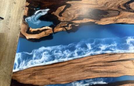 Obere Ansicht einer Tischplatte aus Massivholz mit ausgeprägter Maserung und blau-weißem Epoxidharz-River. Der Tisch steht auf hellem Holzdielenboden neben einer grauen Polsterstuhllehne und wirkt als markanter Blickfang im Raum.