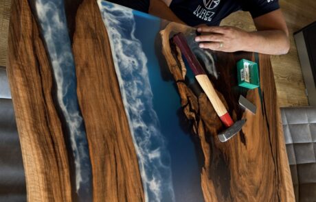 Eine Person in einem JuRez Design T-Shirt präsentiert einen Massivholztisch mit blauem Epoxidharz-River und sichtbarer Baumkante. Werkzeuge liegen auf der polierten Tischplatte, die lebhafte Holzmaserung und eine glänzende Epoxidharzoberfläche zeigt.