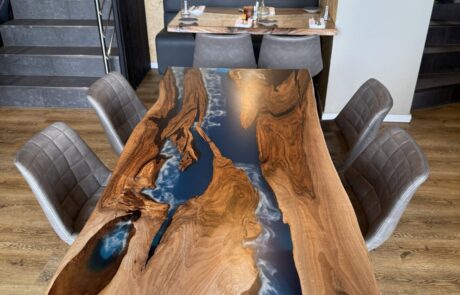 Ein Rivertable aus Massivholz mit tiefblauem Epoxidharz steht in einem modernen Essbereich. Die naturbelassene Baumkante und das schwarze Metallgestell betonen die organische Form und schaffen einen klaren Blickfang.