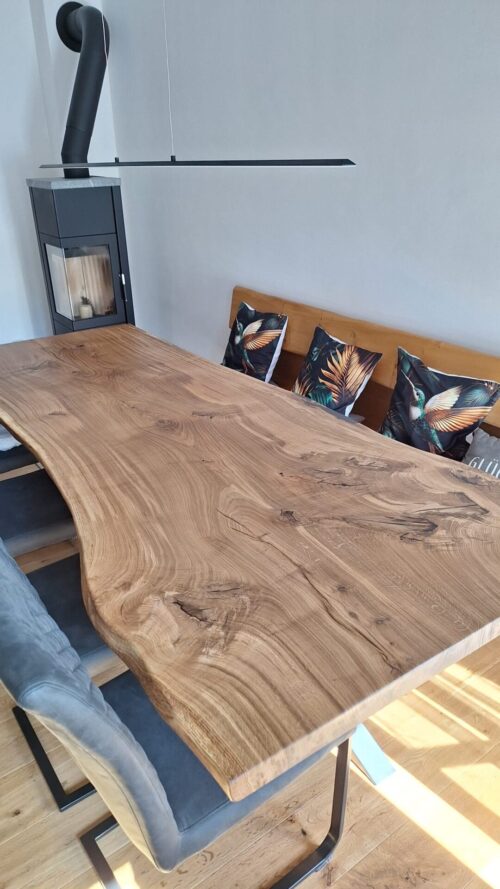 Großer Massivholztisch mit markanter Living Edge Baumkante und sichtbarer Eichenmaserung steht im Essbereich. Ein schwarzes Metallgestell, gepolsterte Stühle, eine Holzbank mit Rückenlehne und ein moderner Holzofen ergänzen die warme Raumwirkung.