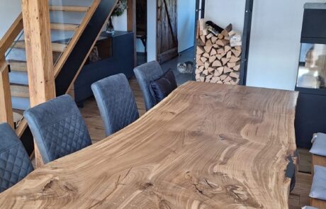 Große massige Echtholz-Tischplatte mit sichtbarer Baumkante und lebhafter Maserung steht auf schwarzem Metallgestell in einem offenen Essbereich. Der Tisch wirkt durch seine natürliche Living Edge und organische Form als zentrales Gestaltungselement im Raum.