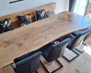 Großer Massivholztisch mit naturbelassener Baumkante und sichtbarer Holzmaserung steht zentral im hellen Essbereich. Eine passende Holzbank und drei dunkelgraue Cantilever-Stühle mit schwarzem Metallgestell rahmen den Tisch und betonen die Verbindung von Naturholz und modernem Design.