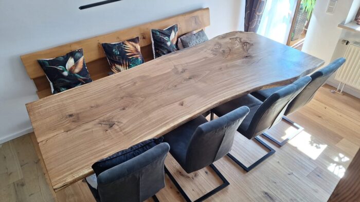 Großer Massivholztisch mit naturbelassener Baumkante und sichtbarer Holzmaserung steht zentral im hellen Essbereich. Eine passende Holzbank und drei dunkelgraue Cantilever-Stühle mit schwarzem Metallgestell rahmen den Tisch und betonen die Verbindung von Naturholz und modernem Design.