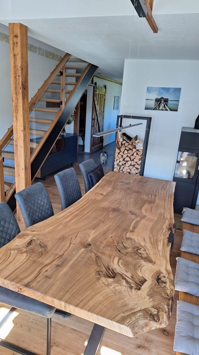 Breite Massivholz-Tischplatte mit markanter Naturkante und ausgeprägter Maserung steht auf einem dunklen Metallgestell im offenen Essbereich. Im Raum sind gepolsterte Stühle, eine Holzbank, ein Holzstapel und eine Treppe mit Holzdetails sichtbar.